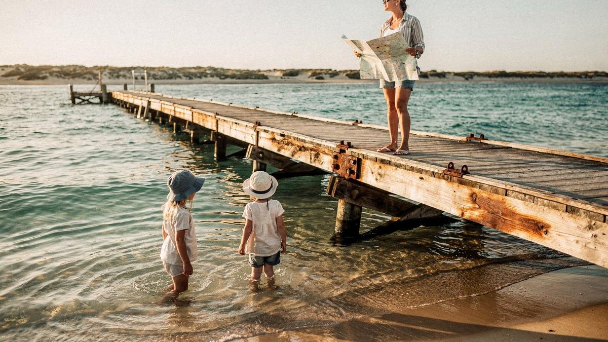 Two children in sun hats wading in shallow clear water beside a wooden jetty while a parent behind them holds a folded map